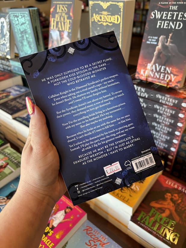 A hand with pink nail polish holding a copy of 'In the Dark' by Shain Rose, featuring silver title text over a dark blue background.