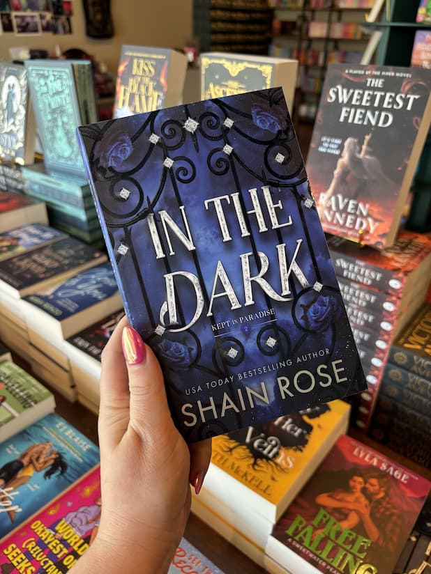 A hand with pink nail polish holding a copy of 'In the Dark' by Shain Rose, featuring large silver title text over a dark blue background with an ornate black wrought-iron gate, blue roses, and small diamond-like accents.