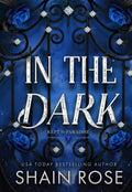 Book cover for 'In the Dark' by Shain Rose, featuring large silver title text over a dark blue background with an ornate black wrought-iron gate, blue roses, and small diamond-like accents.