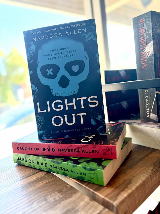 The book 'Lights Out' by Navessa Allen is displayed standing upright on a wooden table, supported by other books including 'Caught Up' and 'Game on'. Each cover features illustrations in blue, red, and green.