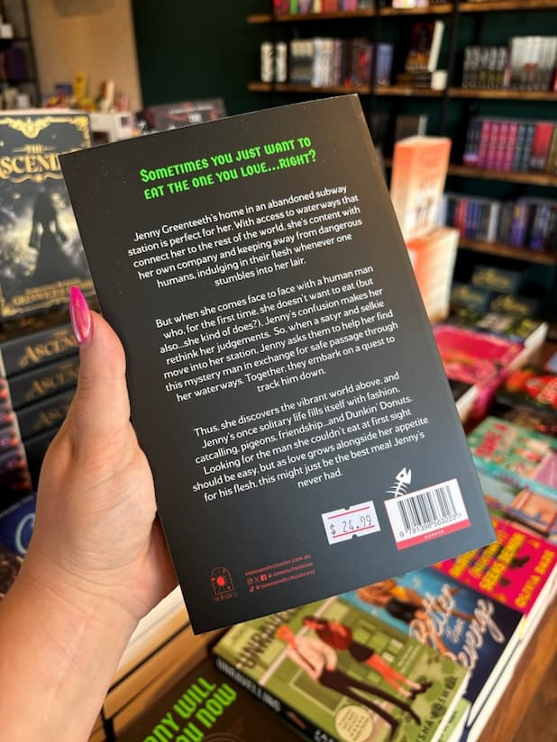 A hand with pink nail polish holding the back of a copy of 'Jenny Will Eat You Now' by Gillian Daniels, featuring a black background and white and green text.