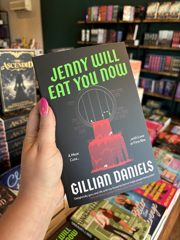 A hand with pink nail polish holding a copy of 'Jenny Will Eat You Now' by Gillian Daniels, featuring a black background, bright green title text, a stylized barred cage, dripping red blood, and white author text.