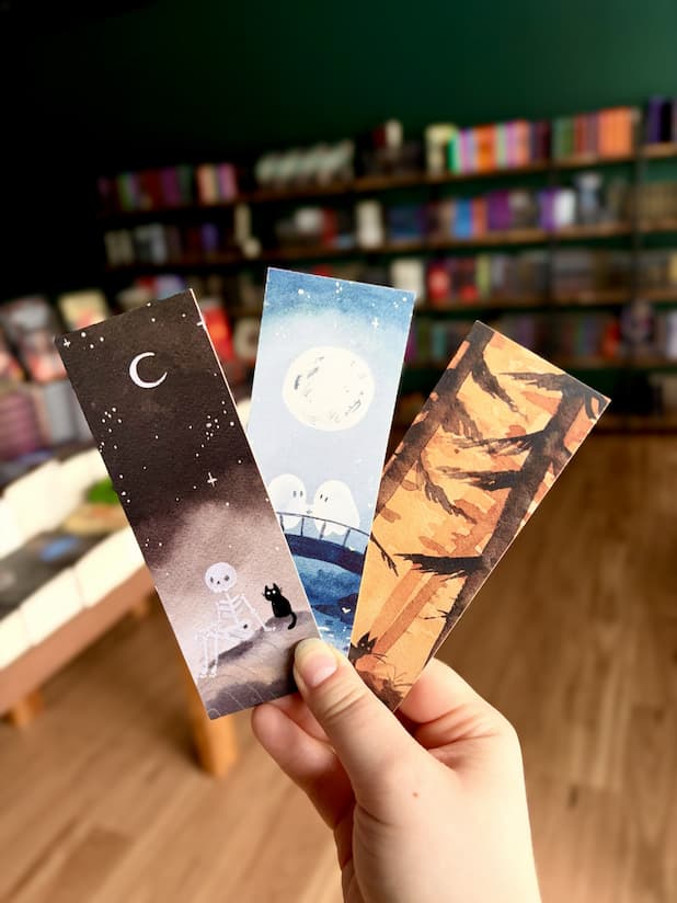 Three illustrated paper bookmarks by Jess Rose are held in a hand in front of a blurred bookstore background with bookshelves and wood flooring. The bookmarks feature whimsical night scenes: a dark starry sky with a crescent moon and small skeleton figure next to a black cat, a pale blue moonlit scene with a bridge and two ghosts, and an orange autumn tree silhouette with a black cat.