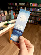 Three illustrated paper bookmarks by Jess Rose are held in a hand in front of a blurred bookstore background with bookshelves and wood flooring. The bookmark features a pale blue moonlit scene with a bridge and two ghosts.