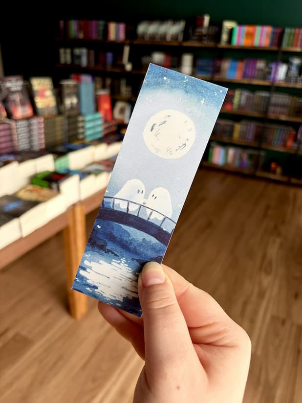 Three illustrated paper bookmarks by Jess Rose are held in a hand in front of a blurred bookstore background with bookshelves and wood flooring. The bookmark features a pale blue moonlit scene with a bridge and two ghosts.