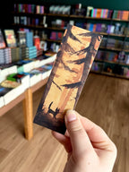 Three illustrated paper bookmarks by Jess Rose are held in a hand in front of a blurred bookstore background with bookshelves and wood flooring. The bookmark features an orange autumn tree silhouette with a black cat.