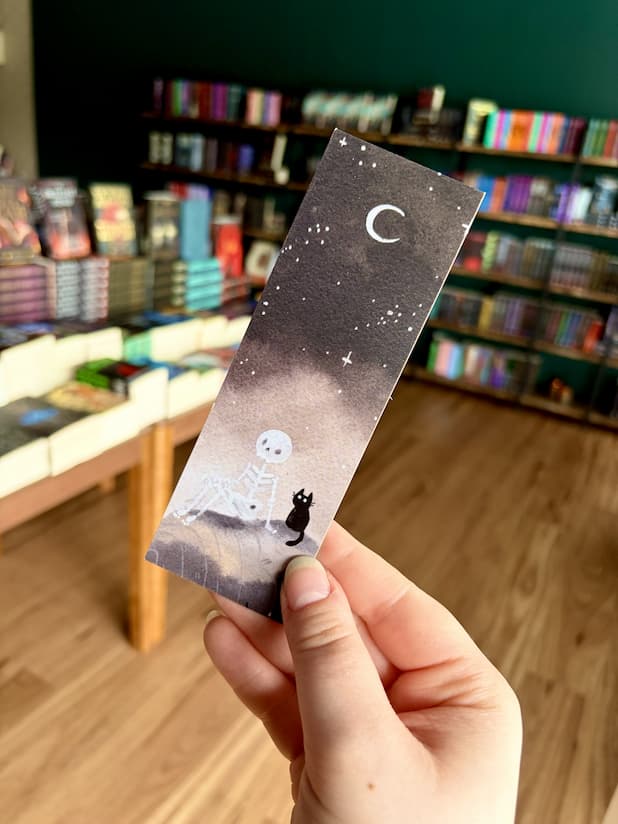 Three illustrated paper bookmarks by Jess Rose are held in a hand in front of a blurred bookstore background with bookshelves and wood flooring. The bookmark features a dark starry sky with a crescent moon and small skeleton figure next to a black cat.