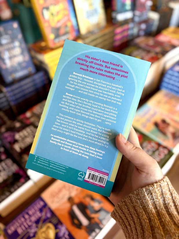 A hand holding the back cover for 'Just for the Plot' by Meghan Quinn, featuring light blue background with white and pink text.