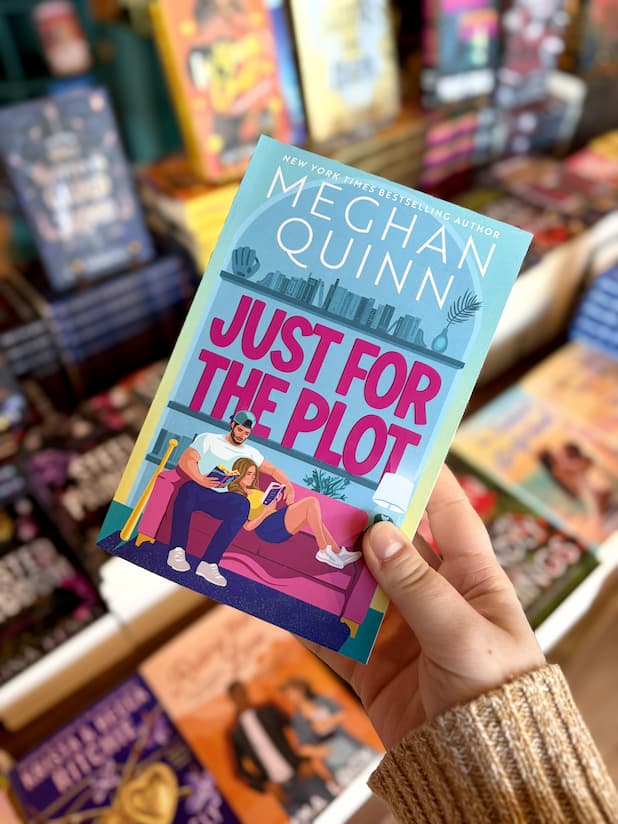 A hand holding a copy of for 'Just for the Plot' by Meghan Quinn, featuring a colorful illustration of a man and woman sitting on a pink couch in a living room, with a woman reclined in his lap. They're reading the same book. 