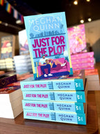 A stack of books for 'Just for the Plot' by Meghan Quinn, featuring a colorful illustration of a man and woman sitting on a pink couch in a living room, with a woman reclined in his lap. They're reading the same book. 