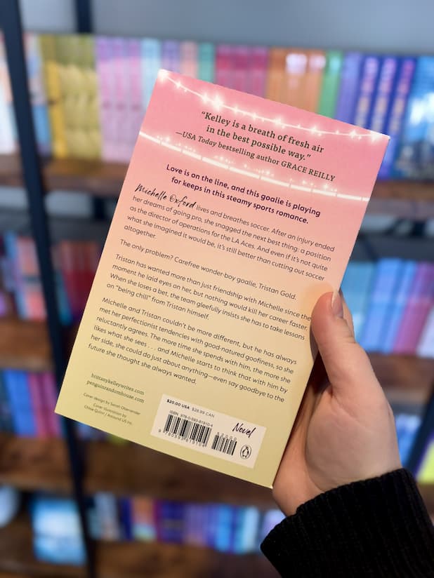 A hand holding the back of a copy of 'Keeping Score' by Brittany Kelley, featuring a pastel yellow and pink gradient and black text.