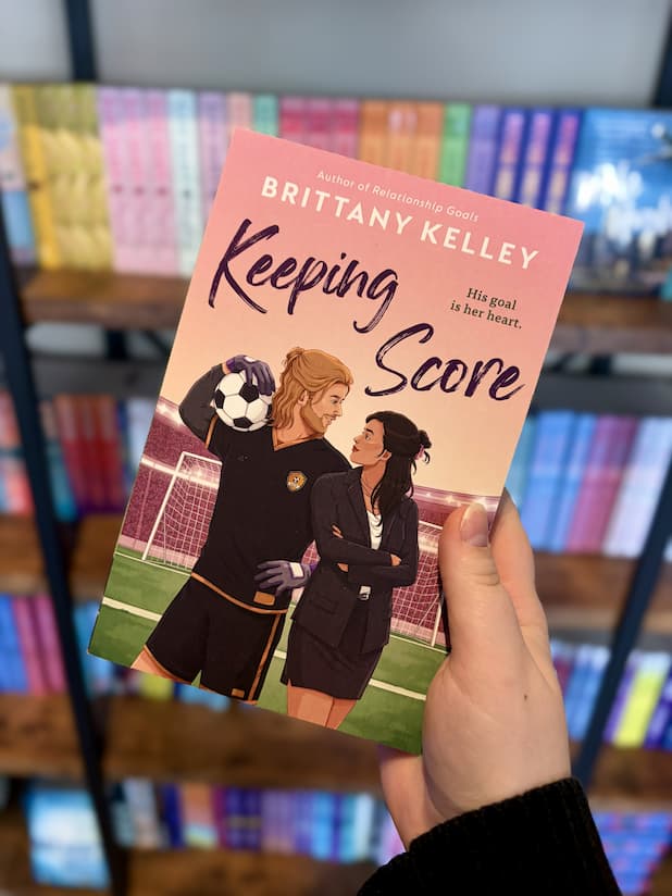 A hand holding a copy of 'Keeping Score' by Brittany Kelley, featuring a soccer player and businesswoman looking across shoulders at each other in front of a stadium.