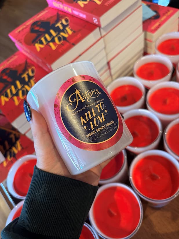 A hand holds a white glass jar candle labeled 'Astoria Kill to Love' in front of stacks of red and pink 'Kill to Love' books and multiple red wax candles in cups.