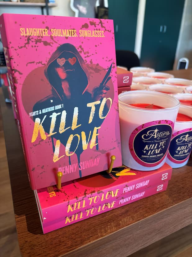 A pink book titled 'Kill to Love' by Penny Sunday stands on a wooden shelf next to several white candles with matching labels. The book cover features a dark hooded figure with heart-shaped sunglasses, and the candles have circular labels with the same title and author branding.