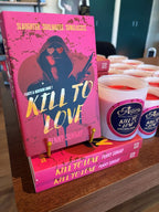 A pink book titled 'Kill to Love' by Penny Sunday stands on a wooden shelf next to several white candles with matching labels. The book cover features a dark hooded figure with heart-shaped sunglasses, and the candles have circular labels with the same title and author branding.
