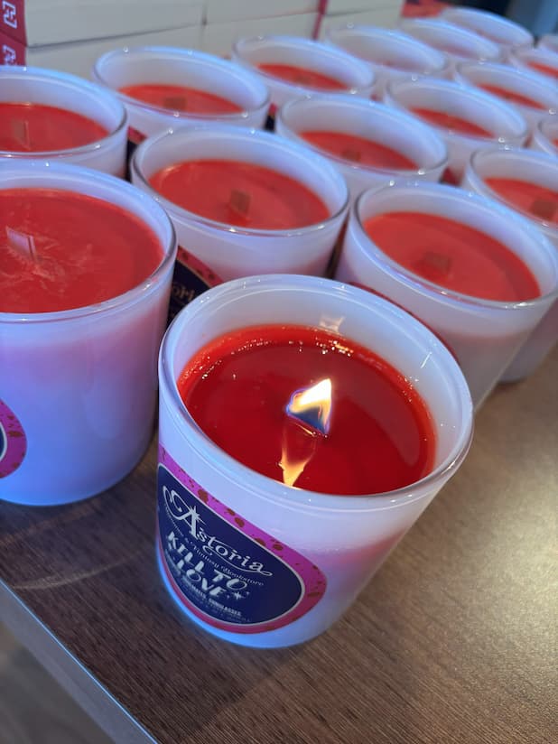 A lit white candle in a clear glass jar with a red wax top, placed on a wooden table alongside multiple unlit candles. The front label reads 'Astoria x Kill to Love'.