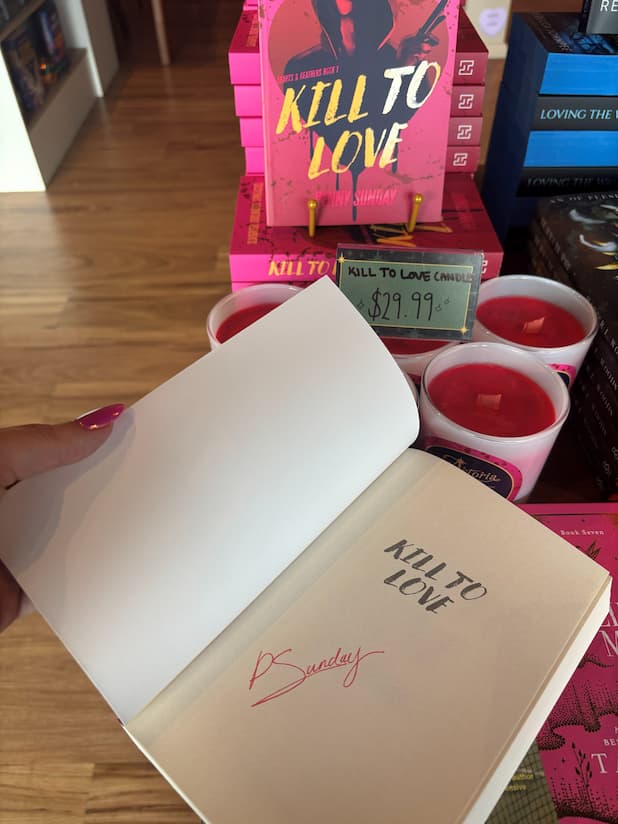 A hand holds an open copy of 'Kill to Love' by Penny Sunday, with a red signature under the title page.