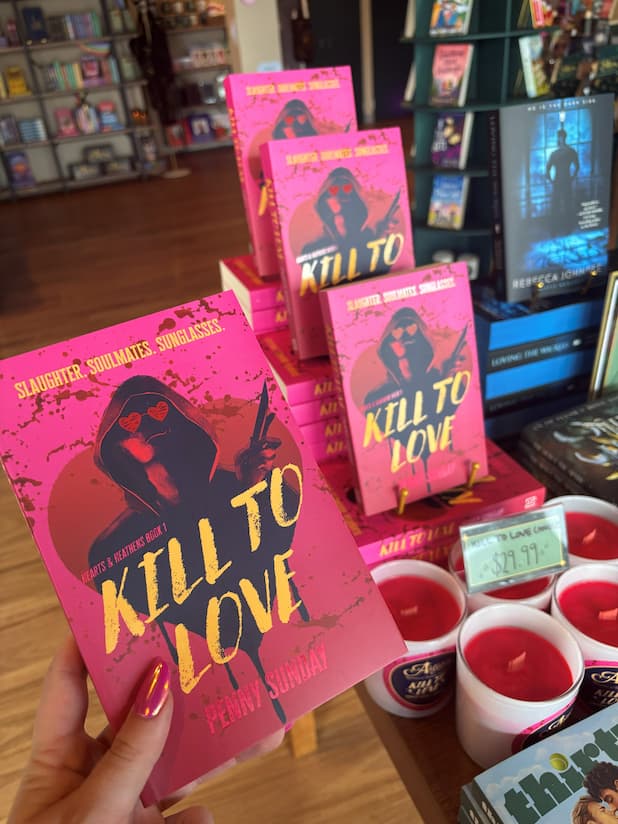 A hand holds the book cover of 'Kill to Love' by Penny Sunday, with a silhouette of a person smirking and wearing red heart-shaped sunglasses on a pink blood splattered background, next to a stack of more copies of the same book.