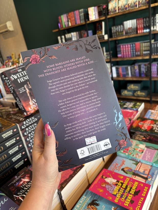 A hand with pink nail polish holding a copy of 'Kiss of the Black Flame' by Helen Scheuerer, featuring a dark purple cover, light pink text, and roses on the corners.