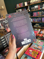 A hand with pink nail polish holding a copy of 'Kiss of the Black Flame' by Helen Scheuerer, featuring a dark purple cover, light pink text, and roses on the corners.