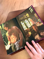 A hand holding open a special edition of 'Kiss of the Black Flame' by Helen Scheuerer, showing the back end pages of an illustrated couple in a fantasy house.