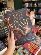 A hand with pink nail polish holding a copy of 'Kiss of the Black Flame' by Helen Scheuerer, featuring an open book surrounded by roses burning from the middle of the book in orange and black flames.