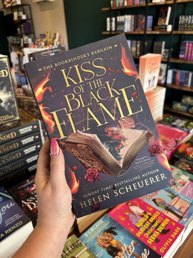 A hand with pink nail polish holding a copy of 'Kiss of the Black Flame' by Helen Scheuerer, featuring an open book surrounded by roses burning from the middle of the book in orange and black flames.