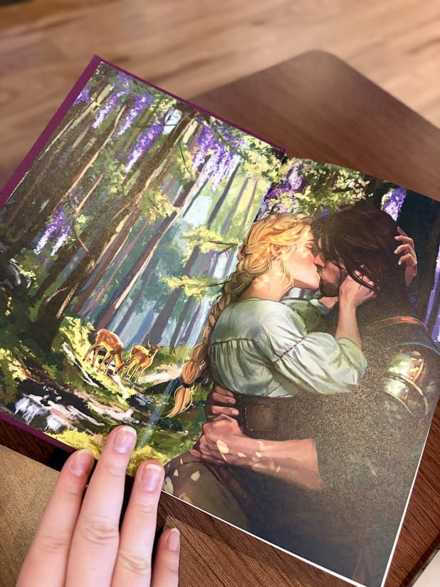 A hand holding open a special edition of 'Kiss of the Black Flame' by Helen Scheuerer, showing the front end pages of an illustrated couple kissing in a forest.