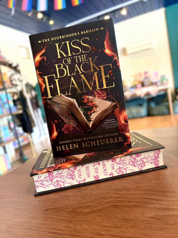 A small stack of the special edition cover of 'Kiss of the Black Flame' by Helen Scheuerer, showing the front cover with a burning open book illustration and patterned pink-and-white sprayed edges.