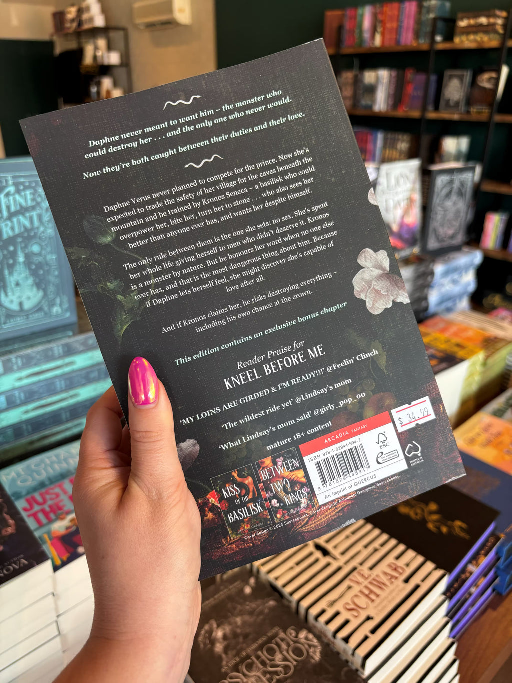 A hand holds the back cover for 'Kneel Before Me' by Lindsay Straube in a bookstore with other books blurry in the background. The cover features a dark background with floral elements, and white text for the blurb.