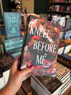 A hand holds the book cover for 'Kneel Before Me' by Lindsay Straube in a bookstore with other books blurry in the background. The cover features a dramatic close-up of a woman with red hair in ornate red clothing, overlaid with large white title text and surrounded by pomegranates, flowers, and metallic decorative elements.
