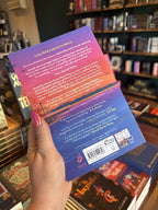 A hand with pink nail polish holds the back of a copy for 'Let's Kiss and Tell' by Joss Richard, featuring a pier with a ferris wheel and a colorful sunset sky.