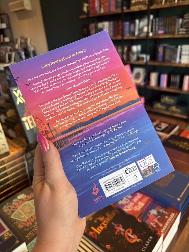 A hand with pink nail polish holds the back of a copy for 'Let's Kiss and Tell' by Joss Richard, featuring a pier with a ferris wheel and a colorful sunset sky.