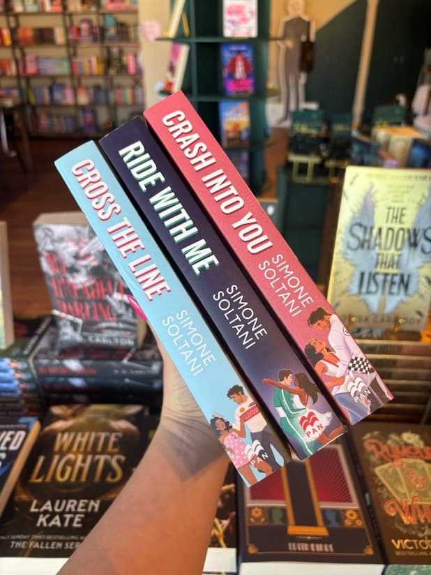 A hand holds three Simone Soltani books in the Lights Out series: Cross the Line, Ride with Me, and Crash Into You. The spines show light blue, dark blue, and coral pink, with white titles and author names.