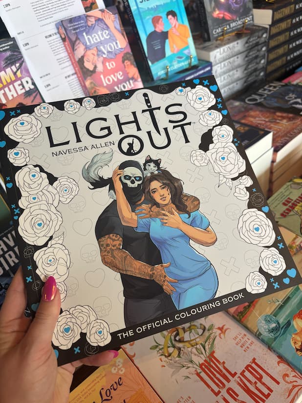 A hand holds the book cover for the Lights Out Official colouring book showing a man in a skull mask embracing a woman in blue scrub with a cat drape over his shoulders in front of a blurry selection of books.