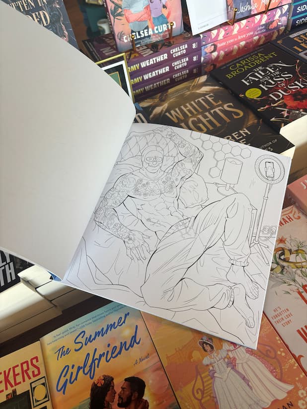 A hand holds the Lights Out Official colouring book open showing an outline of Josh in his mask sitting shirtless, tattoos showing, in sweatpants, on a bed, the book is in front of a table of other books.