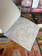 A hand holds the Lights Out Official colouring book open showing an outline of Josh in his mask standing over Aly who is on her knees in front of him, he's holding a knife to her throat, the book is in front of a table of other books.