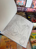 A hand holds the Lights Out Official colouring book open showing an outline of Josh in his mask playing with a cat, the book is in front of a table of other books.