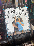 A stack of the Lights Out Official colouring book showing a man in a skull mask embracing a woman in blue scrub with a cat drape over his shoulders in front of a blurry bookstore.