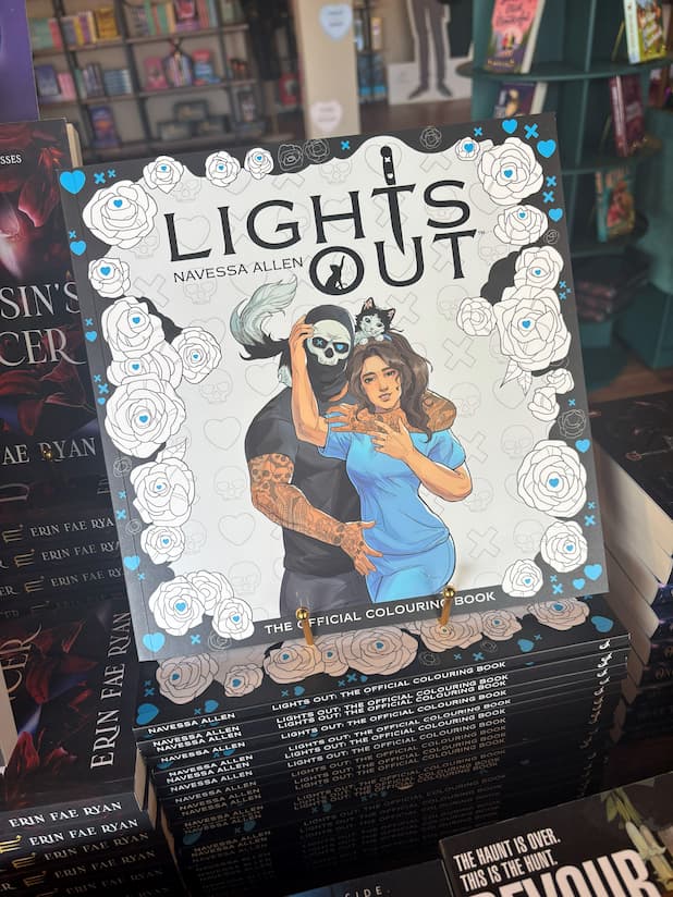 A stack of the Lights Out Official colouring book showing a man in a skull mask embracing a woman in blue scrub with a cat drape over his shoulders in front of a blurry bookstore.