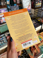 A hand holds the back cover for 'Like in Love With You: A Novel' by Emma R. Alban, featuring purple text on a yellow background with white flower motifs in the corner of the cover, in front of a table full of other blurry books.