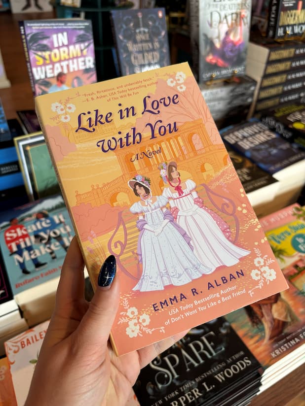 A hand holds the book cover for 'Like in Love With You: A Novel' by Emma R. Alban, featuring an illustration of two women in regency white dresses standing in front of a peach-colored classical building, in front of a table full of other blurry books.