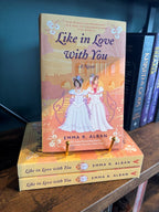 A stack of the book 'Like in Love With You: A Novel' by Emma R. Alban with one book on top showcasing the cover on a gold stand, the cover features an illustration of two women in regency white dresses standing in front of a peach-colored classical building, in front of a table full of other blurry books.