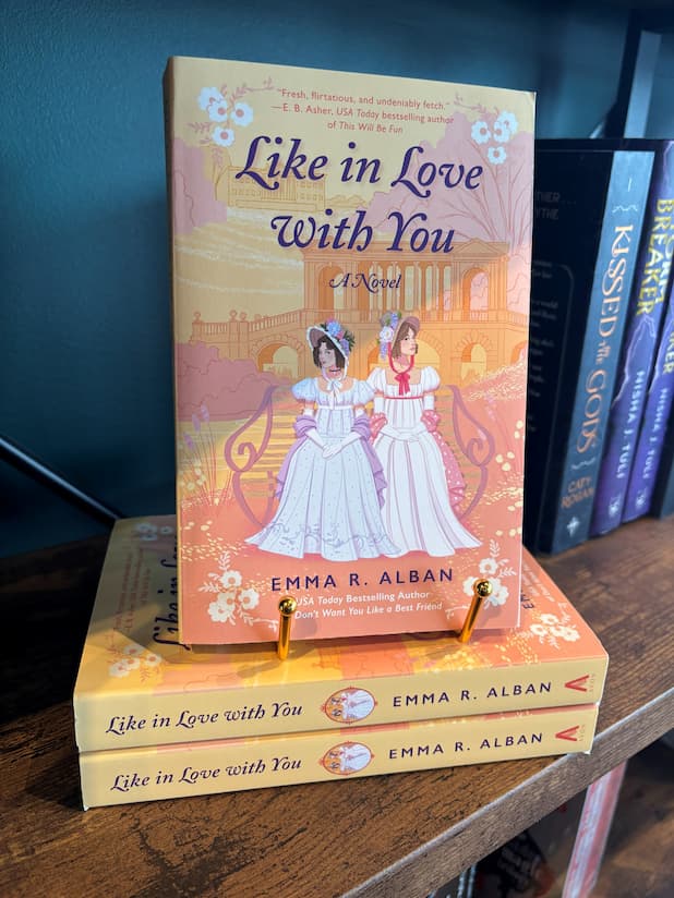 A stack of the book 'Like in Love With You: A Novel' by Emma R. Alban with one book on top showcasing the cover on a gold stand, the cover features an illustration of two women in regency white dresses standing in front of a peach-colored classical building, in front of a table full of other blurry books.