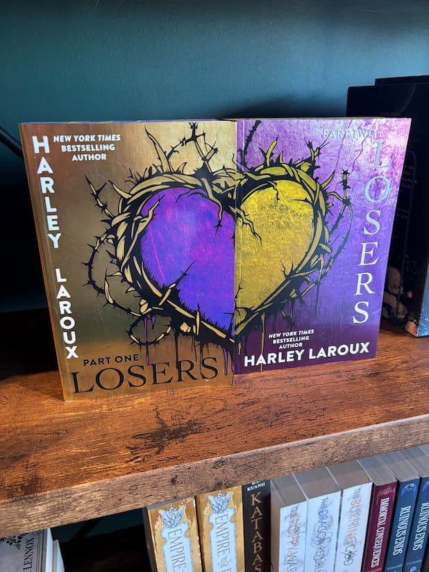 The paperback duet of 'Losers' by Harley Laroux, displayed on a wooden bookshelf. The covers of both books features a split design with a metallic gold and purple mirrored heart surrounded by barbed wire design.