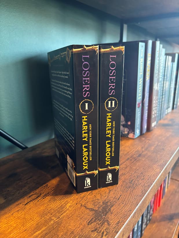 Two paperback books, 'Losers' Volume 1 and 2 by Harley Laroux, standing upright on a wooden bookshelf next to other books.