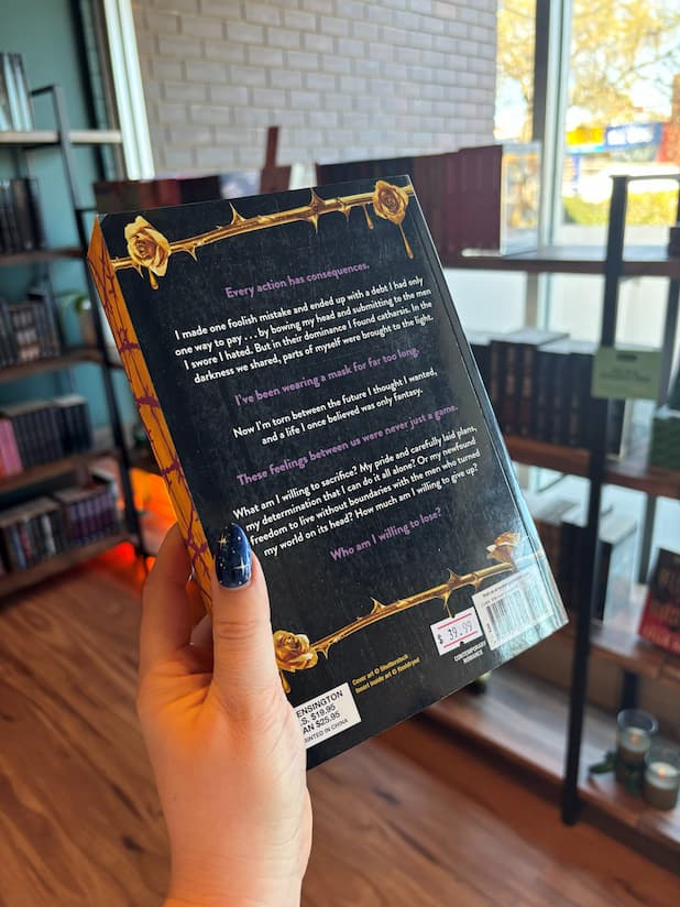 A hand holding the back cover of "Losers: Part Two" by Harley Laroux, with a blurred background of other bookshelves in a bookstore.