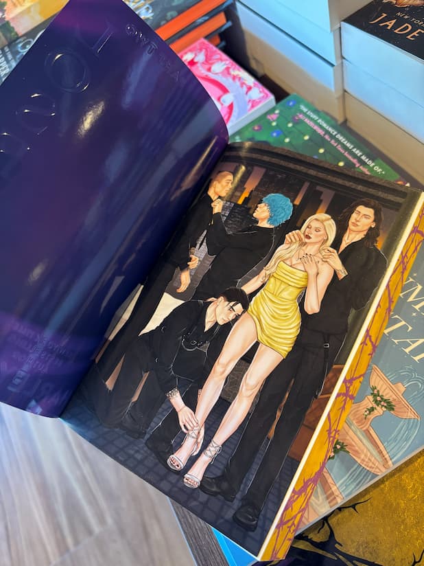 An open copy of "Losers: Part Two" by Harley Laroux, showing an illustration by flashfryed of four men in dark clothing with one woman in a bright yellow dress, they are helping each other get dressed up for an event. The book is photographed indoors with other books and wooden surfaces visible around it.