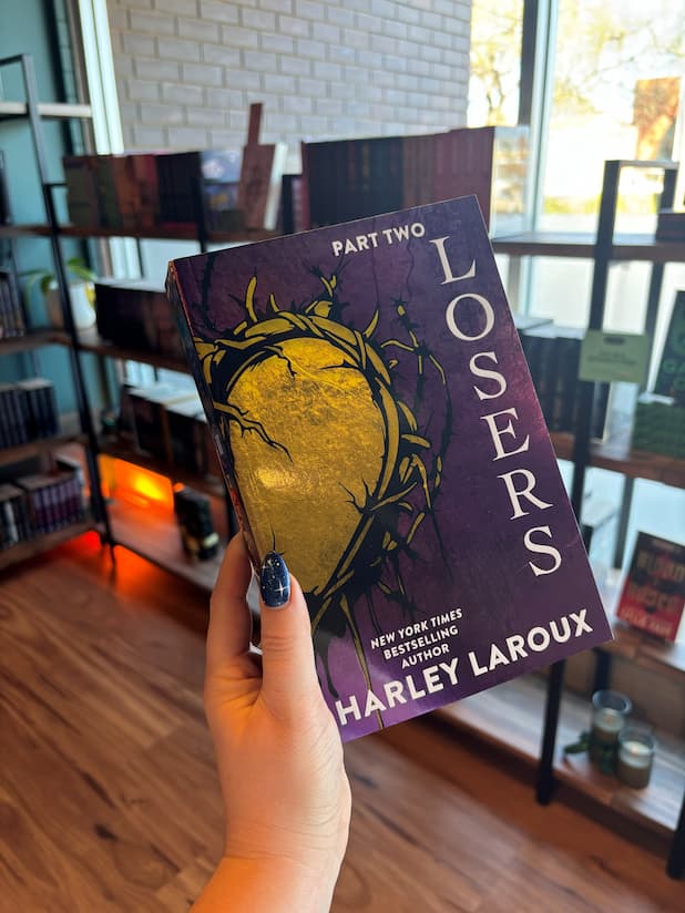 A hand with blue starry nail polish holds a copy of the book 'Losers: Part Two' by Harley Laroux with a metallic purple background and gold barbed wire heart design on the cover and purple and gold sprayed edges.