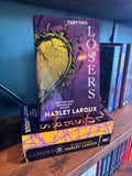 A copy of the book 'Losers: Part Two' by Harley Laroux displayed on a wooden bookshelf, standing upright on a small gold book stand next to other copies of the book.